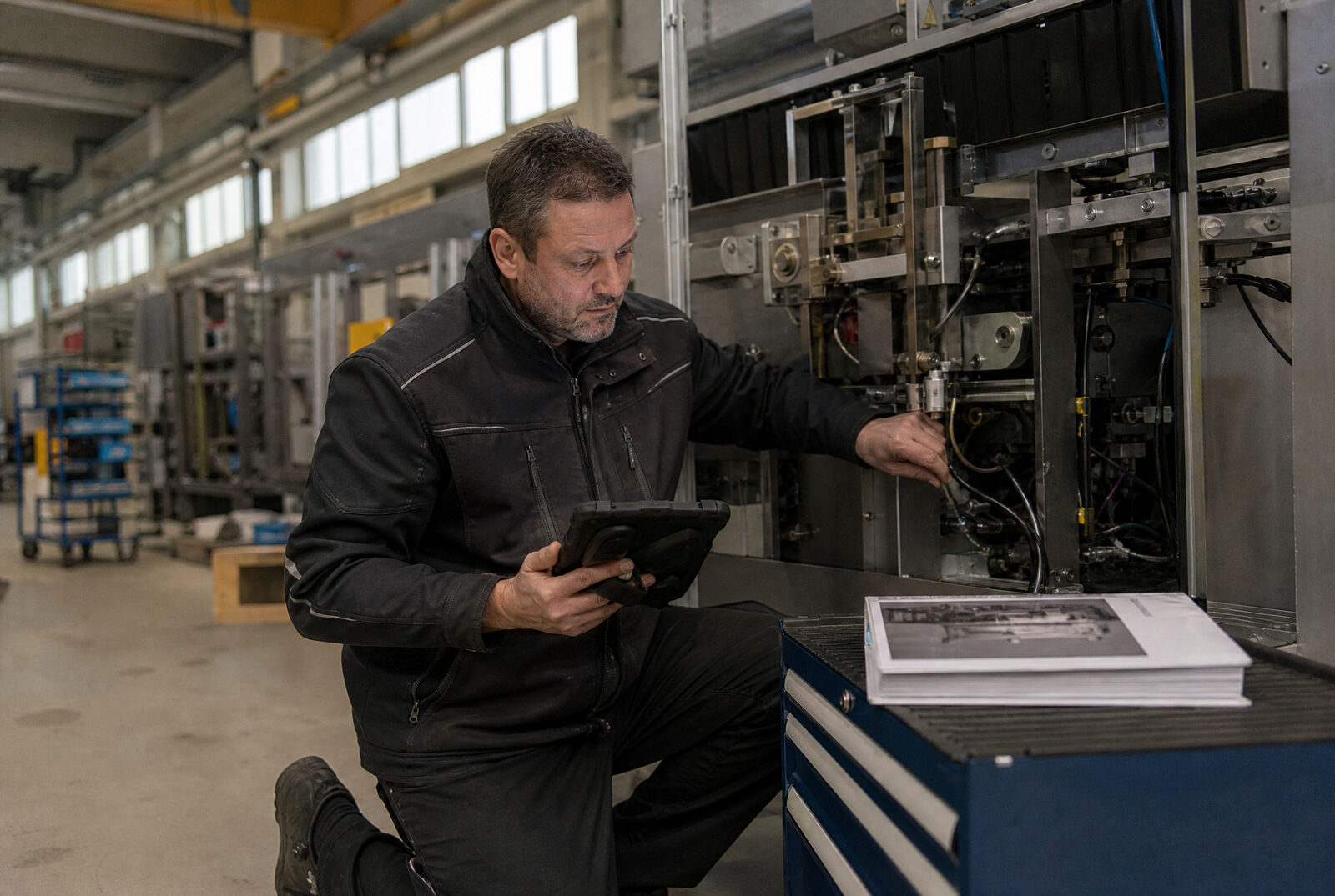 Servicetechniker kniet in einer Produktionshalle an einer geöffneten Maschine und liest auf einem Tablet nach. Auf dem Werkzeugwagen daneben liegt ein Servicehandbuch.